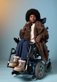 Mladá černošská žena s afro účesom sedí na elektrickom invalidnom vozíku, má na sebe kožušinový kabát, pruhovanú košeľu, tmavomodré nohavice a metalické zimné topánky.