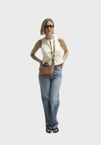 Bruine geweven handtas, beige mouwloze top met strikdetail, lichtblauwe high-waisted jeans en zwarte sandalen. Model staat met de handen op de heupen.