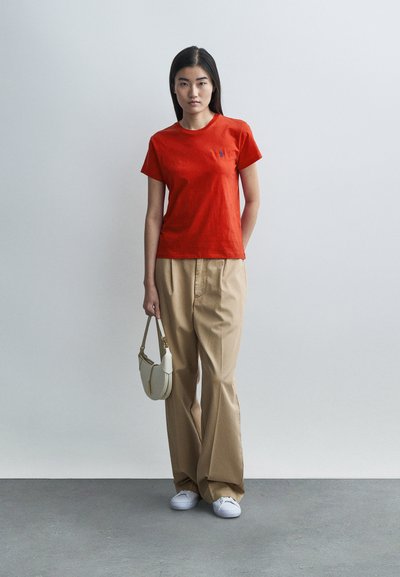 Piros pamut rövid ujjú póló kék logóval, bézs bő szárú nadrággal és fehér sportcipővel, kezében egy kis világos színű kézitáskával.