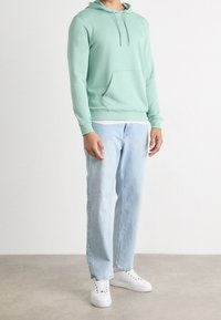 Mátově zelená mikina s kapucí a klokaní kapsou, doplněná světle modrými džíny s rovnými nohavicemi a bílými teniskami na neutrálním pozadí.