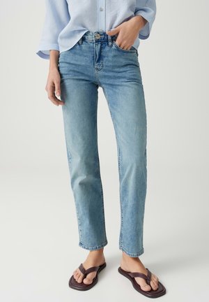 Person, die hellblaue Jeans und ein hellblaues Hemd mit Knöpfen trägt, steht mit einer Hand in der Tasche und braunen Flip-Flops an den Füßen.