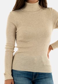 Beige gerippter Rollkragenpullover mit langen Ärmeln. Hat eine taillierte Passform und Knopfdetails an den Manschetten. Weicher Stoff.