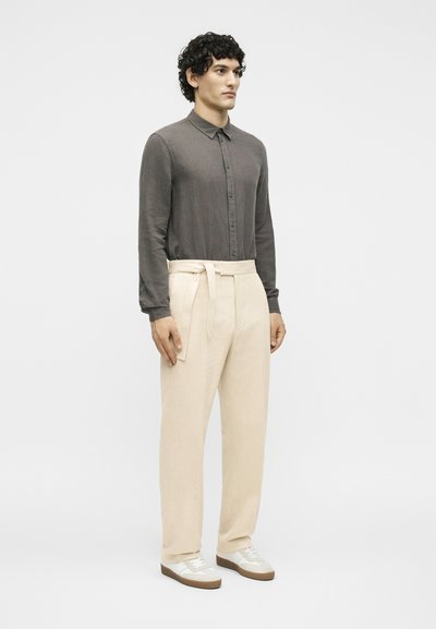 Άνδρας με σγουρά μαλλιά που φοράει σκουρόγριχο πουκάμισο με κουμπιά, μπεζ ψηλόμεσο παντελόνι με δεμένη ζώνη και λευκά αθλητικά παπούτσια, στεκόμενος με πρόσωπο προς τα εμπρός.