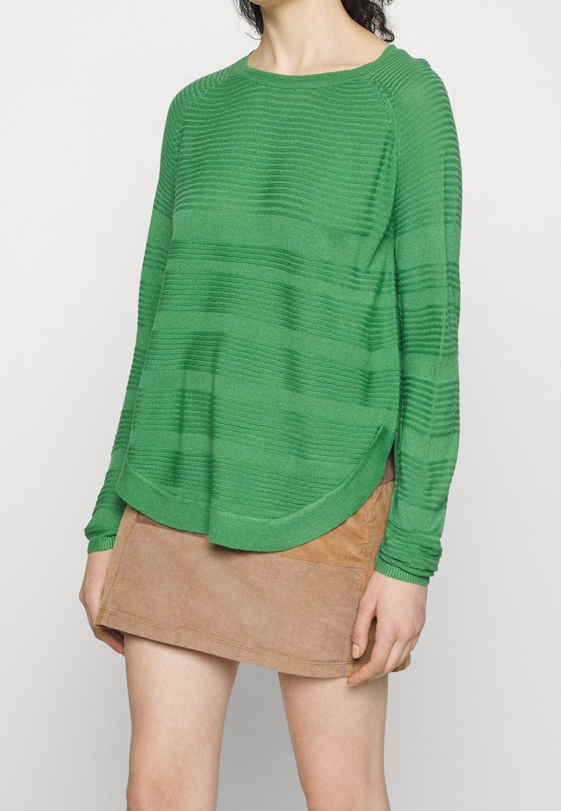 Femme portant un pull texturé vert à manches longues et une mini jupe beige, debout devant un fond clair uni.
