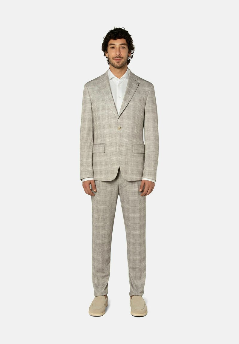 Mann in beigem kariertem Anzug, weißem Hemd und beigen Slip-On-Schuhen, der vor einem schlichten hellen Hintergrund steht.