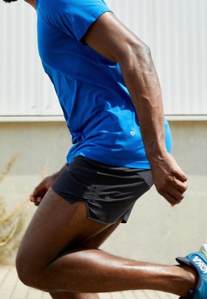 Läufer mit blauem Shirt, schwarzen Shorts und blauen Turnschuhen, der beim Laufen auf einer befestigten Oberfläche im Freien aufgenommen wurde.
