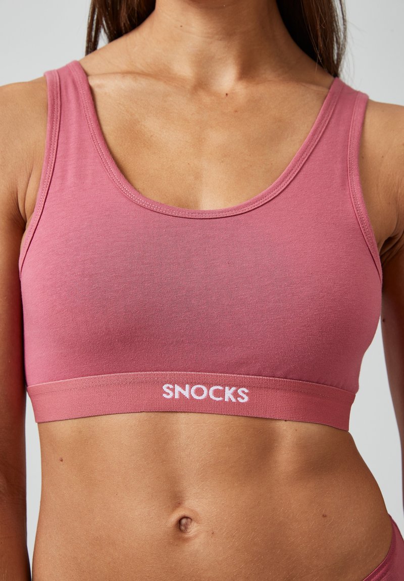 Roze naadloze sportbeha van zachte stof, met een ronde halslijn en een brede band met het "SNOCKS" logo in het wit gedrukt.