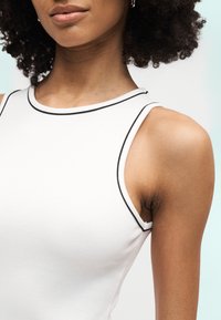 Κοντινή λήψη ενός ατόμου που φορά λευκό αμάνικο τοπ με μαύρο περίγραμμα, παρουσιάζοντας τον ώμο, τον αυχένα και τα χείλη του σε φωτεινό φόντο.