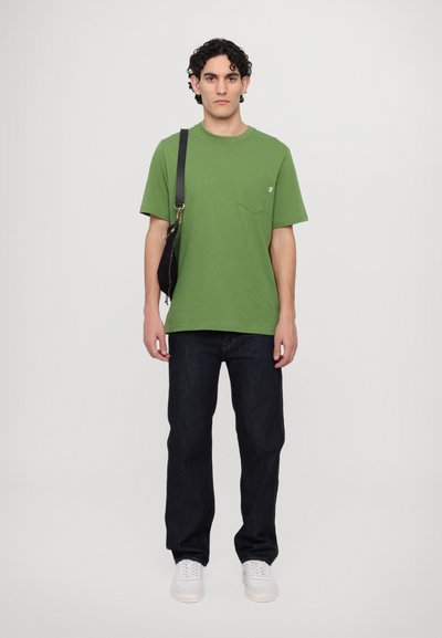 Green short-sleeve t-shirt with a chest pocket, paired with dark blue jeans and white sneakers. Model holding a black crossbody bag.