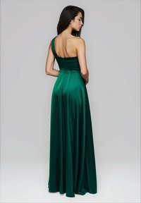 Donna con lunghi capelli scuri che indossa un abito in satin verde smeraldo lungo fino a terra, con una spallina, in piedi davanti a uno sfondo grigio chiaro uniforme.