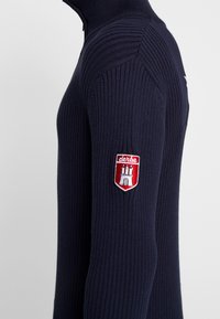 Marinblå stickad tröja med hög krage, ribbad struktur, lång ärm och en röd broderad patch på vänster arm.