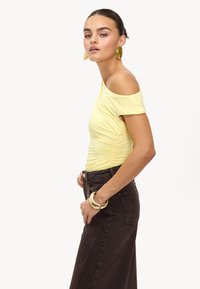 Haut jaune à épaules dénudées avec une texture froncée, associé à une jupe longue taille haute marron foncé. Accentué par de grandes boucles d'oreilles en or.