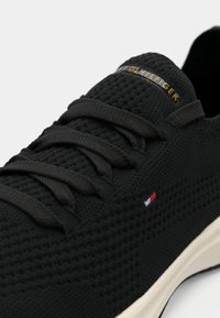 Black knit sneakers with textured upper, featuring flat black laces and a gold logo. A small tricolor accent is visible on the side.