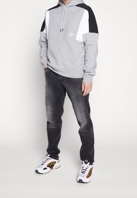 Huppari harmaana, jossa on valkoisia ja mustia yksityiskohtia, nyörillinen huppu, yhdistettynä tummanharmaisiin farkkuihin ja valkoisiin tennareihin, joissa on punaisia ja keltaisia yksityiskohtia.