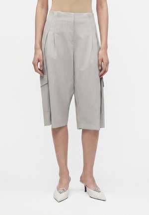 Kobieta nosząca jasnoszare, szerokie krótkie spodenki sięgające do kolan z bocznymi kieszeniami oraz białe, spiczaste mules na wysokim obcasie z ozdobami.