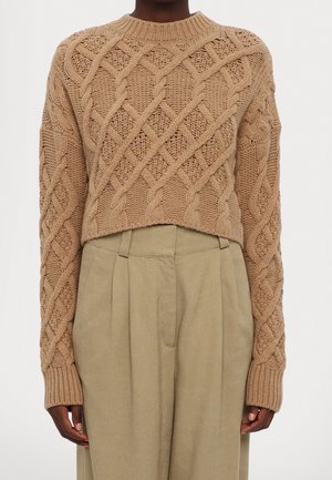 Personne portant un pull court en grosse maille beige avec un motif torsadé et un pantalon ample beige taille haute, debout devant un fond uni.