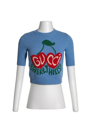 Niebieski krótki dzianinowy crop top z czerwonym wzorem wiśni i zielonym tekstem "GUCCI BEVERLY HILLS" z przodu, prezentowany na białym torsie manekina.
