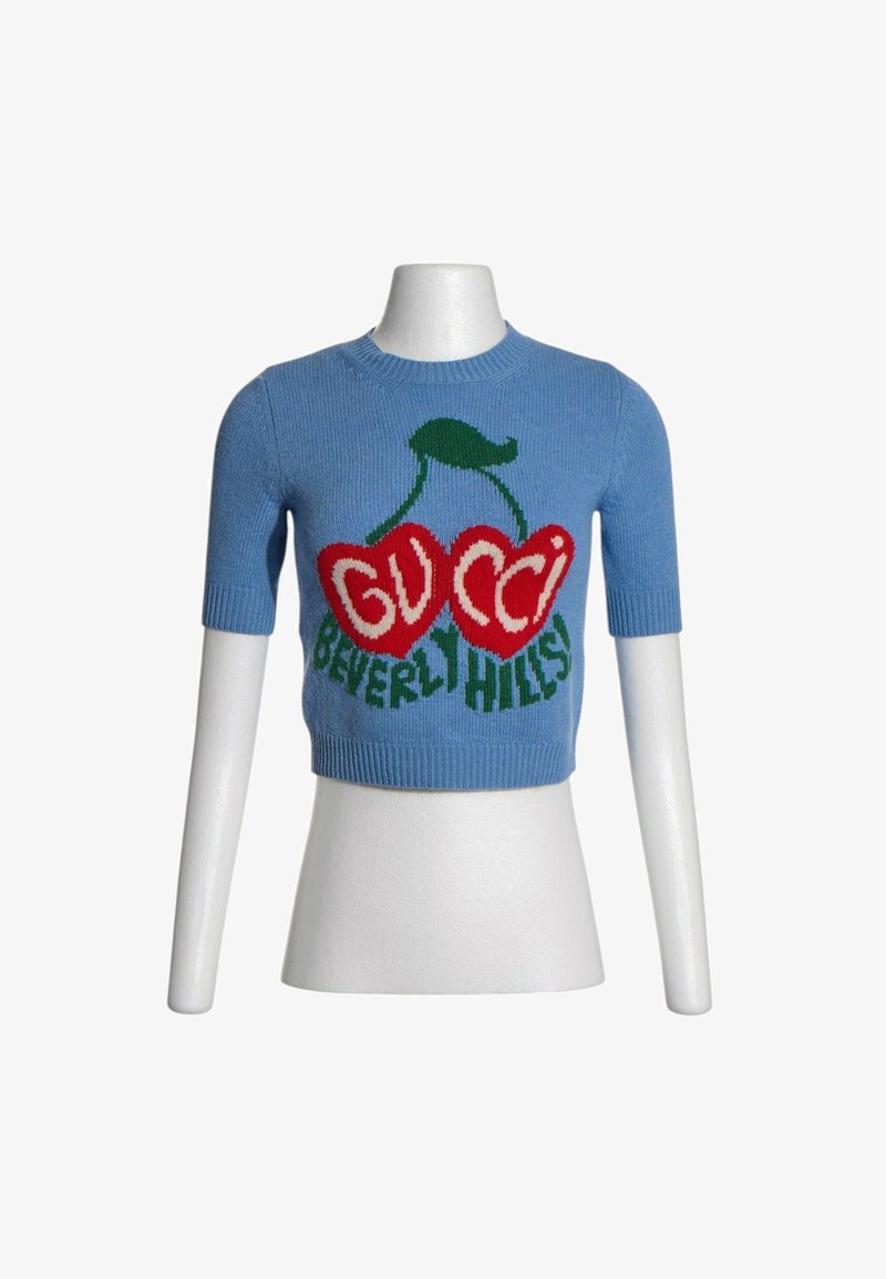 Crop top tricoté bleu à manches courtes avec motif cerise rouge et texte vert "GUCCI BEVERLY HILLS" à l'avant, présenté sur un torse de mannequin blanc.