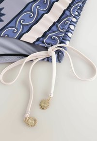 Gros plan sur un bas de bikini à motif bleu avec des liens latéraux blancs réglables se terminant par des breloques dorées en forme de soleil.