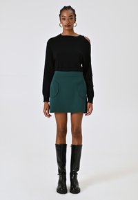 Maglione nero con aperture sulle spalle, abbinato a una mini gonna verde con tasche tonde e una texture liscia. Stivali neri al ginocchio.