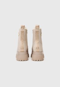 Pár béžových suedových kotníkových bot s tlustými lugovými podrážkami, vertikálními poutky se značkou a viditelnou ovčí vlnou uvnitř.