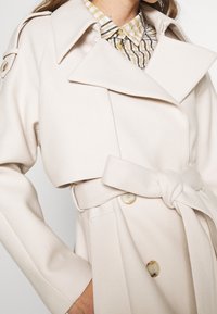 Beżowy płaszcz trench z szerokimi klapami, pasek w talii i podwójnym zapięciem na guziki. Posiada teksturowaną tkaninę i usztywniony kołnierz.