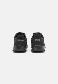 Chaussures de sport noires avec une tige en maille, un collier rembourré et le logo JAKO sur le talon. Elles possèdent une semelle en caoutchouc texturée pour une meilleure traction.