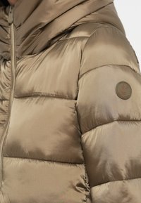 Gedeelte pufferjack in matte beige stof met gequiltte secties. Heeft een capuchon en een ronde logo patch op de linkermouw. Ritssluiting.