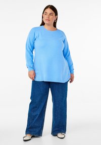 Helles blaues, langärmliges Sweatshirt mit rundem Halsausschnitt, weicher Textur und lockerer Passform, kombiniert mit dunkelblauen Schlagjeans und schwarz-weißen Schuhen.