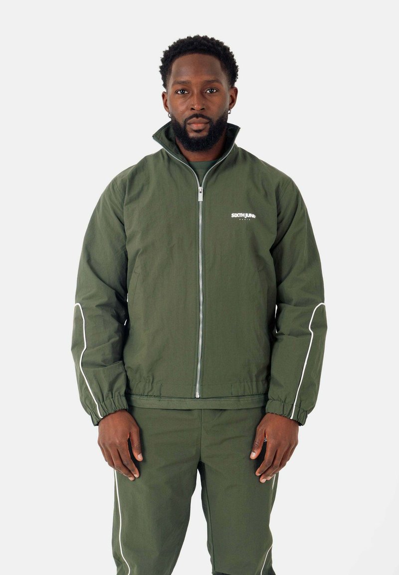 Olijfgroene jas met hoge kraag en witte accenten, voorzien van een rits aan de voorkant, elastische manchetten en een logo op de borst. Gecombineerd met bijpassende broeken.