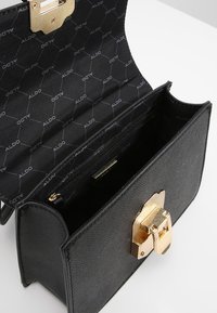 Czarna torebka o fakturowanej powierzchni z złotym zatrzaskiem, wnętrze wyścielone czarną podszewką z powtarzającym się wzorem logo, wyposażona w kieszeń na suwak.