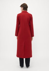 Person wearing a long, red coat with a back slit, standing facing away, paired with black pants and black heeled shoes.
