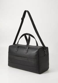 Black duffle bag made of textured leather. Features two handles, a detachable shoulder strap, and a horizontal fabric band across the front.