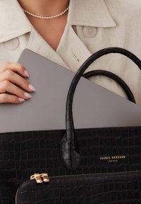 Schwarze Handtasche mit Krokodilprägung, zwei Griffen, goldenen Reißverschlussakzenten und Branding. Hält einen grauen Laptop in der Nähe eines beigen Kleidungsstücks.