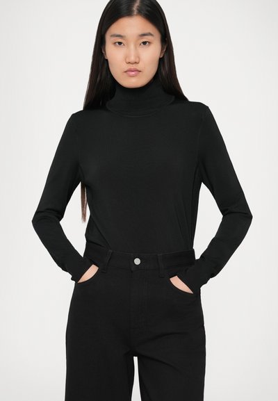 Top negru cu guler înalt, cu mâneci lungi, design ajustat, textură rib, purtat cu pantaloni negri cu talie înaltă, cu mâinile în buzunare.