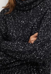 Pull à col roulé en maille mouchetée noir et blanc, manches longues, texture douce, coupe décontractée, avec un col et des poignets roulés.