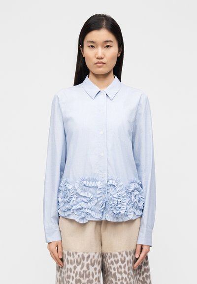 Chemise à rayures bleu clair avec un motif floral à volants à l'ourlet. Comprend un col et des manches longues, portée avec un pantalon en velours côtelé beige.