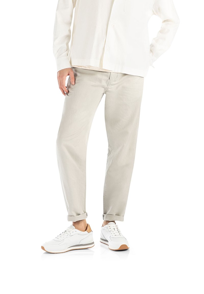 Personne portant un pantalon beige clair avec les ourlets retroussés et des baskets blanches avec des accents marron, debout sur un fond blanc.