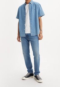 Světle modrá džínová košile s krátkým rukávem přes bílé tričko, kombinovaná se světle modrými džínami a černými teniskami s bílými podrážkami.