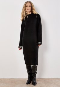 Schwarzes Strickkleid mit langen Ärmeln, weißen gestreiften Bündchen und geripptem Saum. Kombiniert mit schwarzen kniehohen Stiefeln.