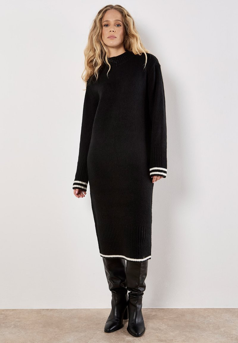 Schwarzes Strickkleid mit langen Ärmeln, weißen gestreiften Bündchen und geripptem Saum. Kombiniert mit schwarzen kniehohen Stiefeln.