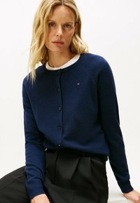 Cardigan azul marino de material suave, con cuello redondo, cierre de botones y un pequeño detalle de logo. Lleva puesto sobre una camisa blanca.