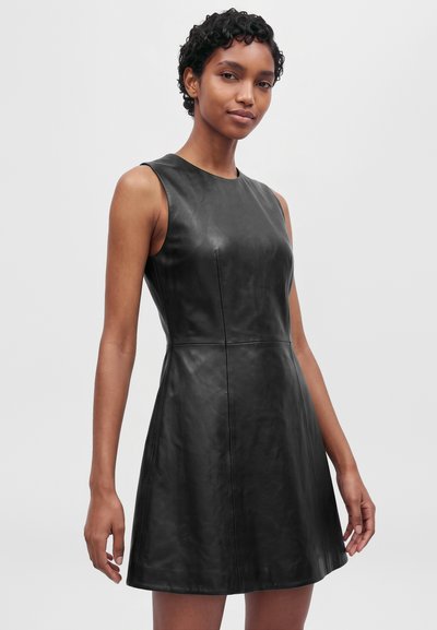 Un modèle aux cheveux courts et bouclés pose devant un fond uni, vêtu d’une robe noire sans manches d’aspect cuir, ajustée à la taille et évasée dans le bas.