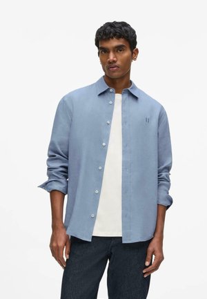 Junger Mann mit lockigem schwarzem Haar, der ein hellblaues Hemd mit Knöpfen über einem weißen T-Shirt und dunklen Jeans trägt, steht vor einem schlichten weißen Hintergrund.