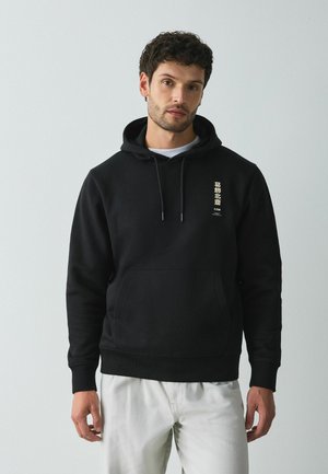 Mann mit kurzen lockigen Haaren, der einen schwarzen Hoodie mit vertikalem weißen Text auf der Brust und helle Hosen vor einem einfarbigen Hintergrund trägt.