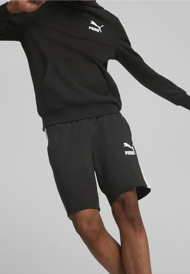 Felpa nera Puma con tasca frontale, abbinata a pantaloni corti neri. La felpa presenta un logo bianco; i pantaloni corti hanno strisce bianche e un logo.