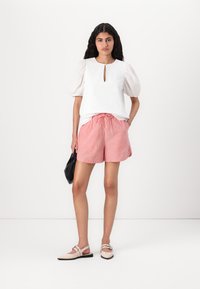 Haut blanc texturé avec manches bouffantes et détail en forme de clé, associé à un short à rayures rouges et blanches avec cordons de serrage et des ballerines de couleur claire.