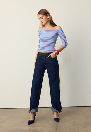 Ung kvinde med langt hår iført en blå off-shoulder stribet top, mørke opsmøgede vidde jeans, høje hæle og farverige armbånd.