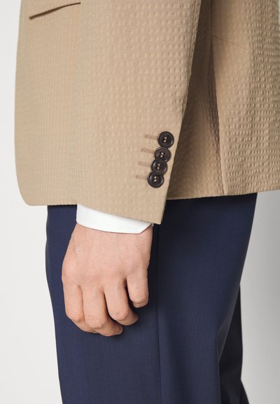 Manga de um blazer texturizado bege com quatro botões escuros, usado sobre uma camisa branca e calças escuras, destacando o corte e os detalhes sob medida.