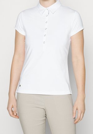 Vrouw draagt een wit poloshirt met korte mouwen met knoopsluiting en beige broek, staand met ontspannen armen langs het lichaam.
